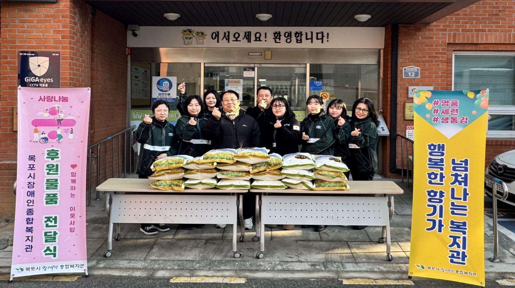 목포영성교회 관계자들이 목포시장애인종합복지관 현관 앞에서 쌀 10kg 20포대를 후원하고 기념촬영을 하고 있다. 복지관 입구 자동문 상단에는 '어서오세요! 환영합니다!'라는 문구가 적혀 있다. 입구 앞 테이블 위에는 쌀 포대들이 차곡차곡 쌓여 있고, 검은색 점퍼를 맞춰 입은 9명의 교회 관계자들이 쌀 뒤에 서서 손가락 하트 포즈를 취하며 미소 짓고 있다. 사진 양옆에는 복지관의 후원 전달식 안내 배너와 홍보 배너가 각각 설치되어 있다.